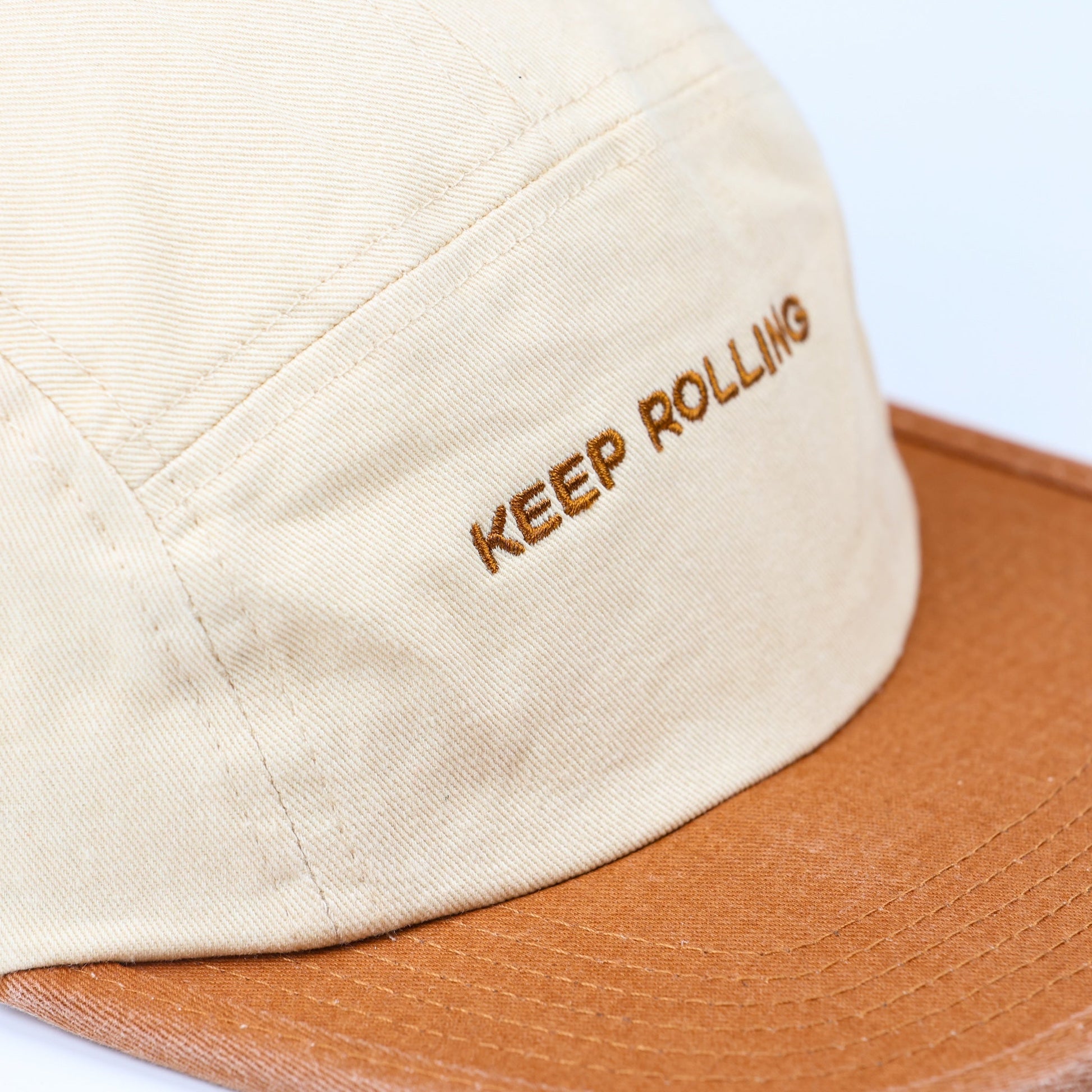 Close-up of a beige 5-panel cap with a brown leather-look flat brim and brown embroidered text reading 'KEEP ROLLING' on the side, set against a plain white background.