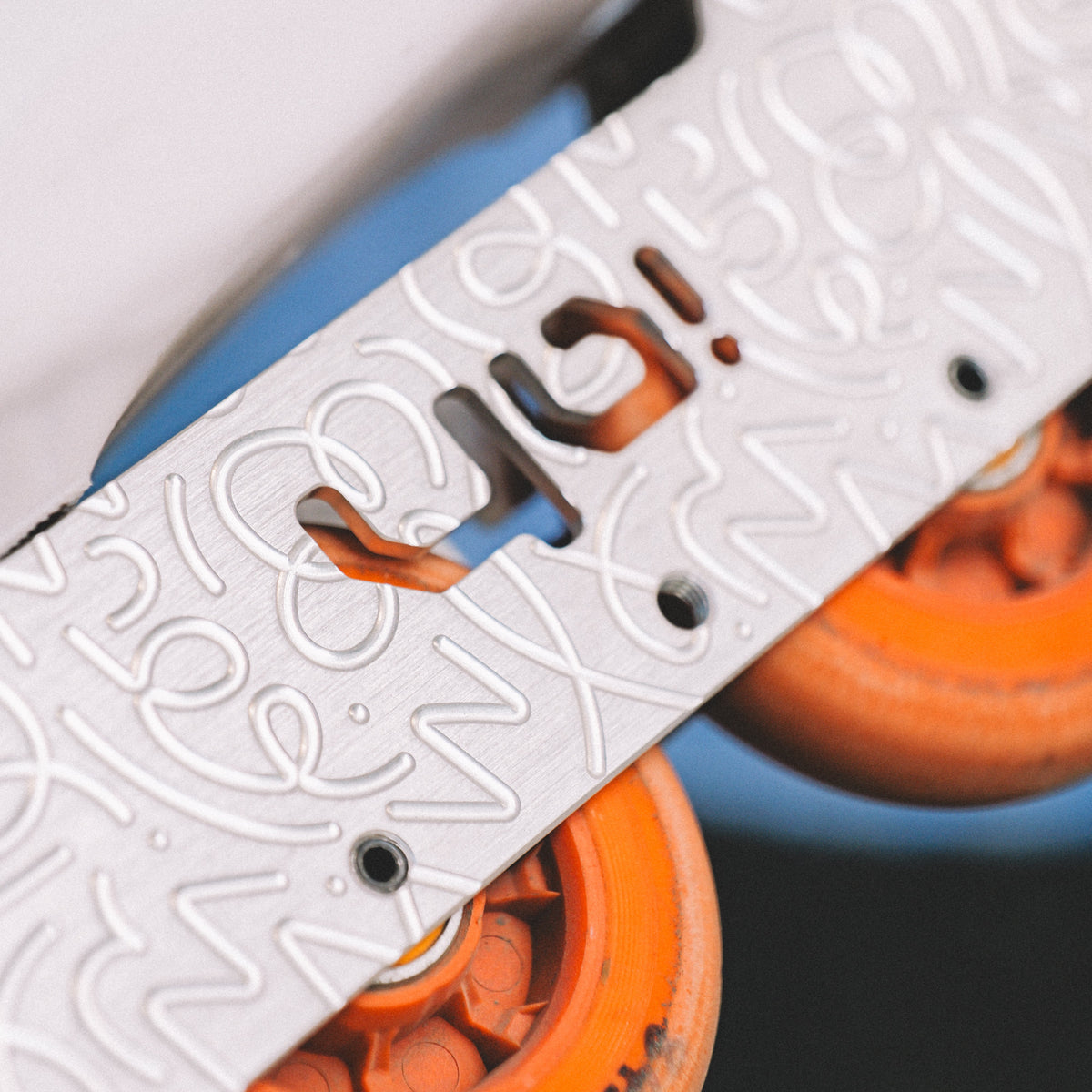 Close-up of a white inline skate with orange wheels and a white metal plate featuring a stylized cutout logo.