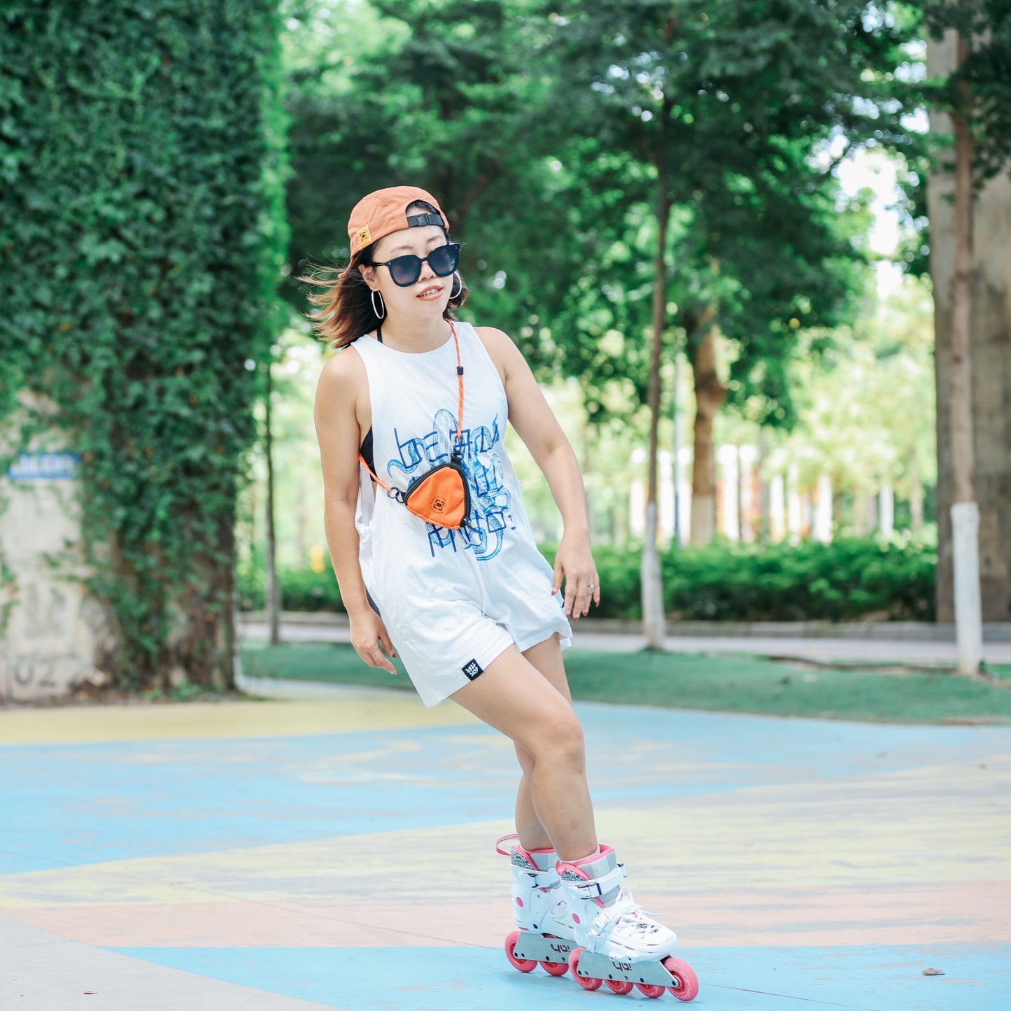 a  girl wearing YOYOSKATE  Capsule Pouch  with a blue graphic, white shorts, a brown cap, and sunglasses is inline skating outdoors on a colorful paved path with a green ivy-covered wall in the background.