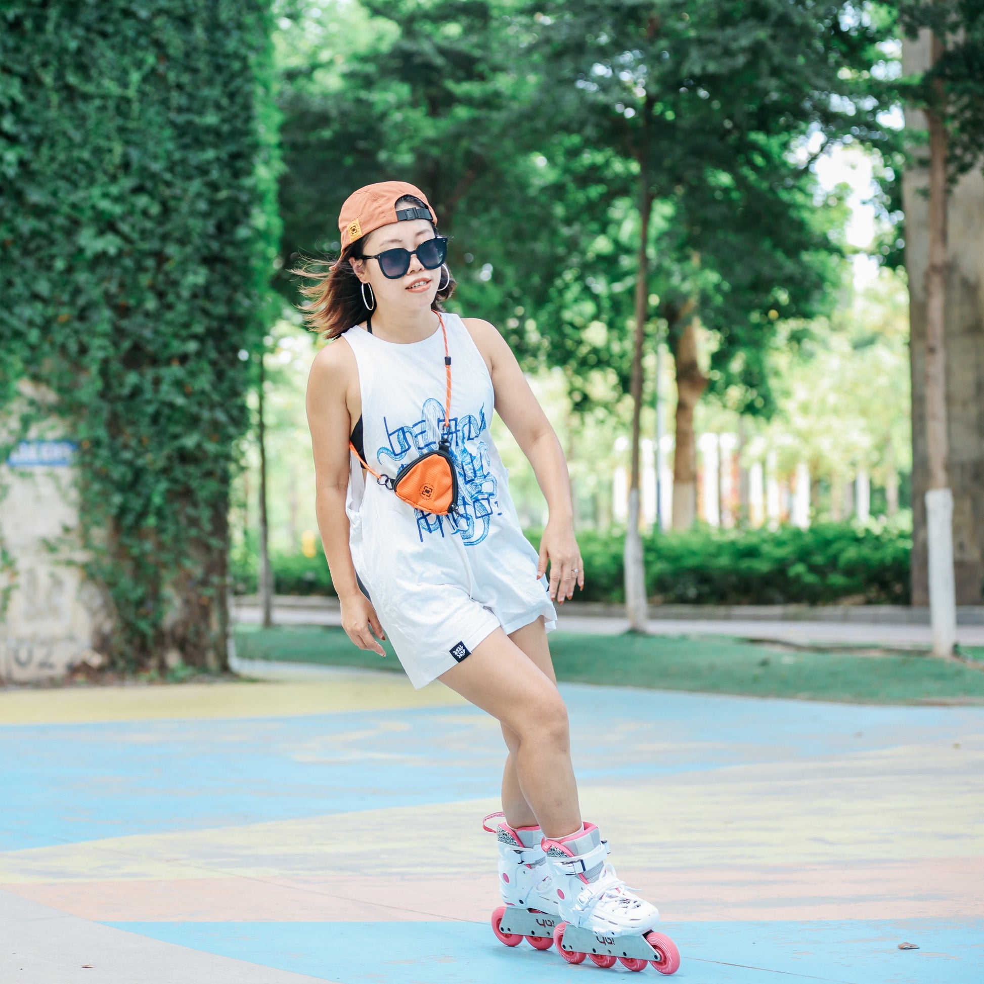 a  girl wearing YOYOSKATE  Capsule Pouch  with a blue graphic, white shorts, a brown cap, and sunglasses is inline skating outdoors on a colorful paved path with a green ivy-covered wall in the background.