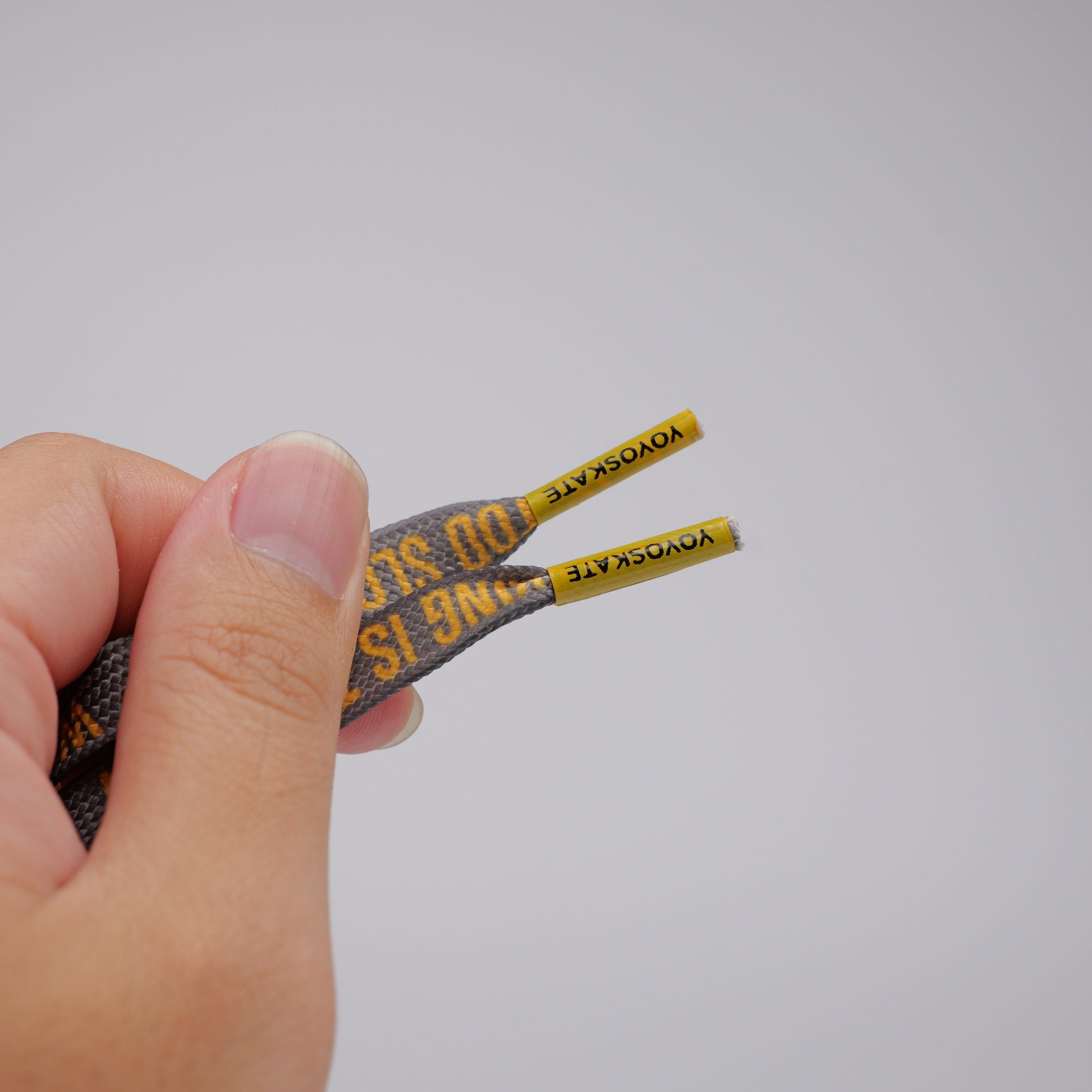 A close-up of a hand holding a pair of shoelaces with a dark gray and yellow patterned woven design and yellow end tabs against a plain light gray background.