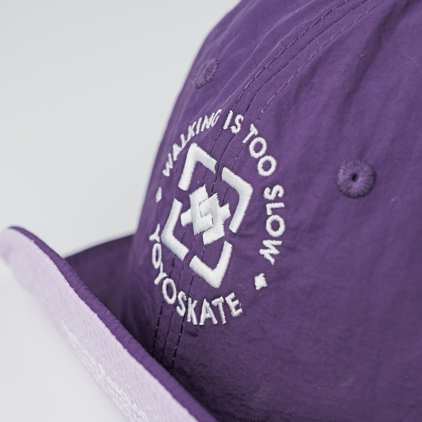 A close-up of a purple snapback cap featuring a white embroidered logo and the text 'WALKING IS TOO SLOW @YOSKATE' on the side panel.