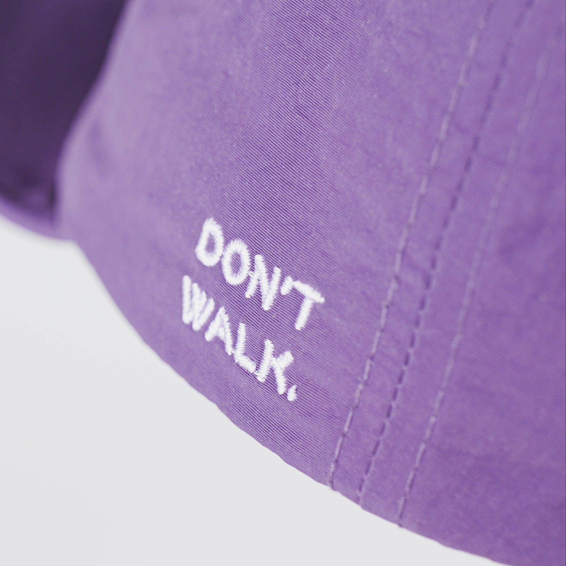 Close-up of a purple baseball cap with white embroidered text reading 'DON'T WALK.' on a light gray background.