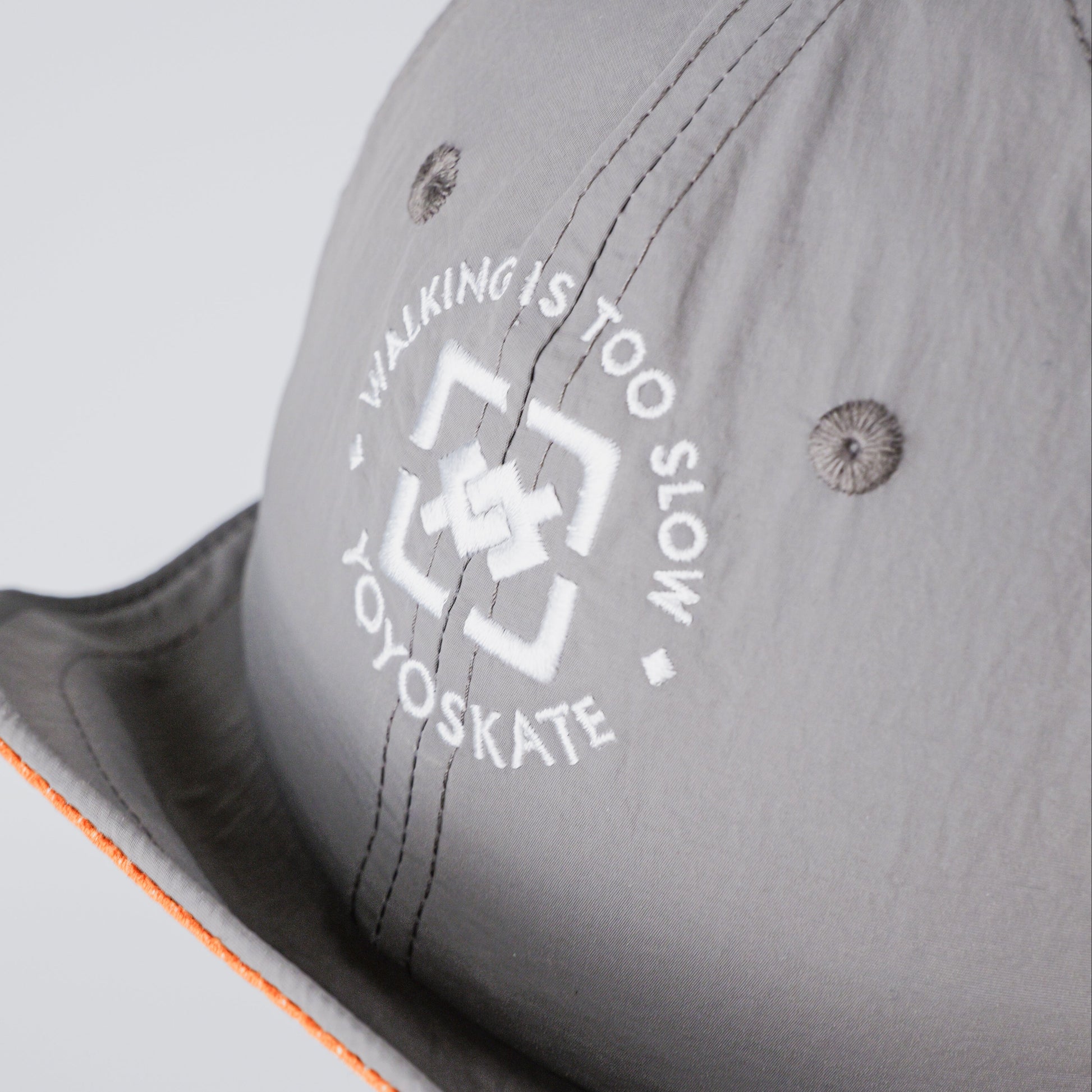 Close-up of a light gray wide-brim cap with a white circular 'Walking is too slow Oyoskate' logo on the front panel and a bright orange lining visible along the brim edge.