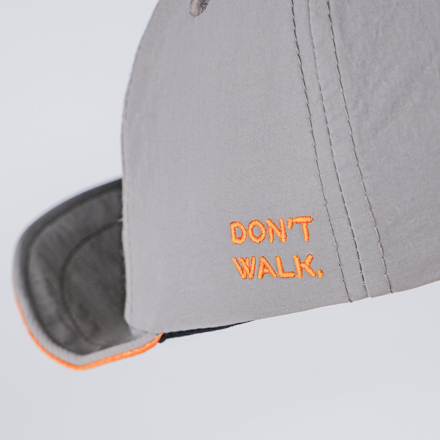 A close-up of a light gray baseball cap with orange embroidered text reading 'DON'T WALK.' on the side, shown against a plain light gray background.