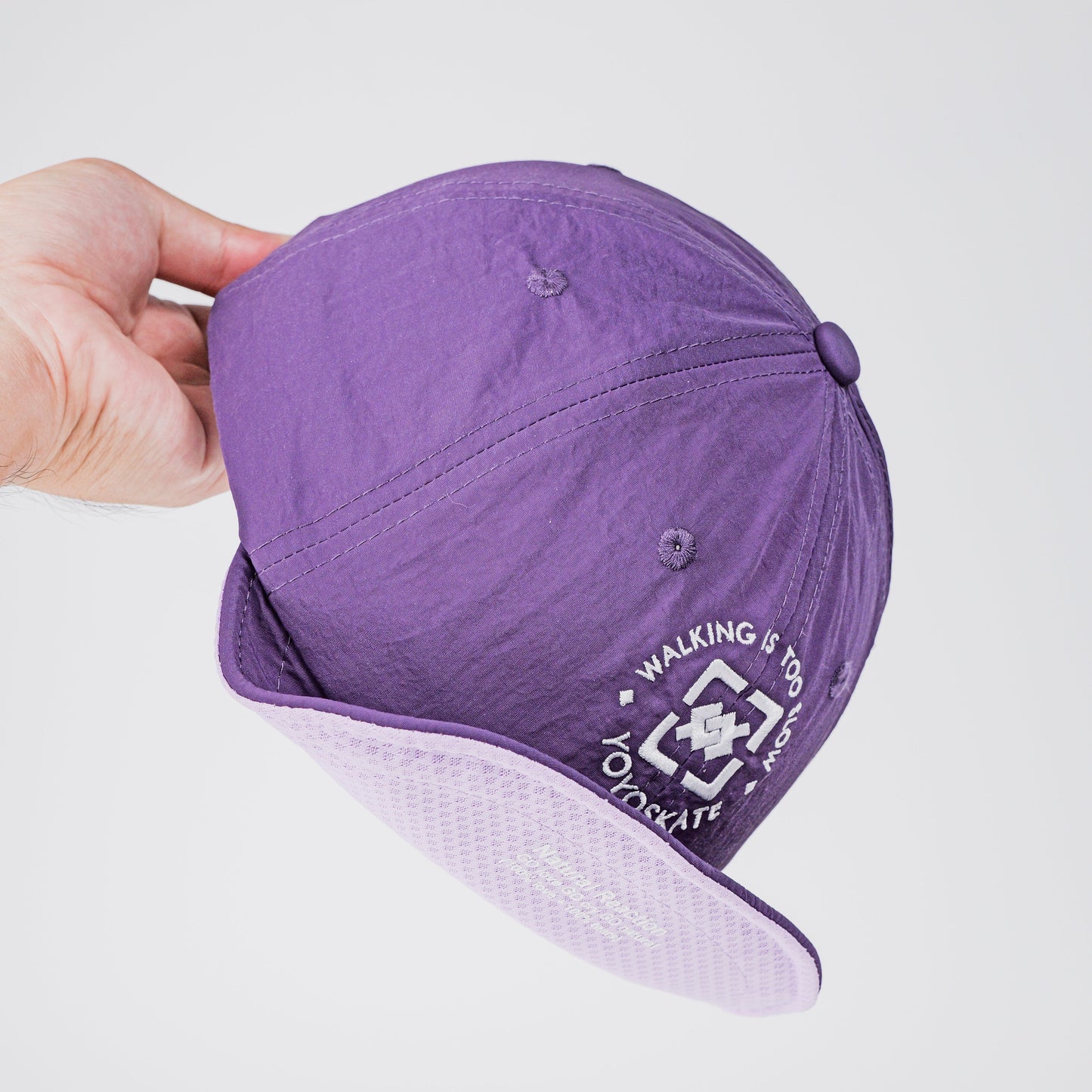 A purple baseball cap held by a hand against a light gray background, showing a white embroidered logo on the side and a light lavender underside on the brim.