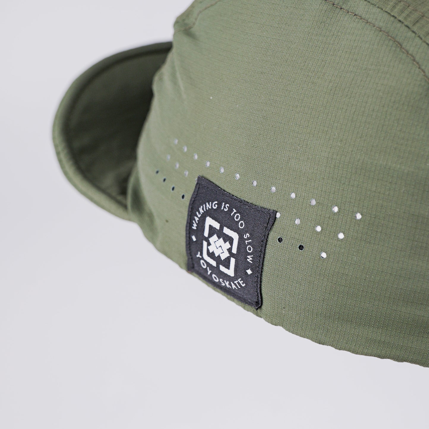 Close-up of an olive green cap with a black brand patch on the side and small white dot details, photographed against a light gray background.