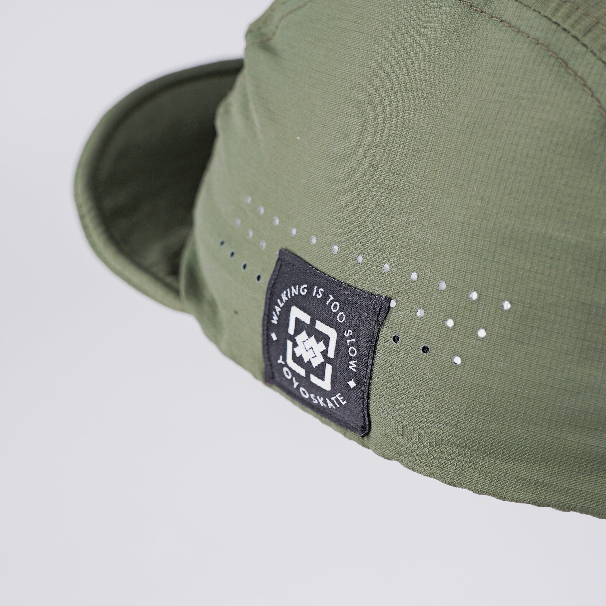 Close-up of an olive green cap with a black brand patch on the side and small white dot details, photographed against a light gray background.