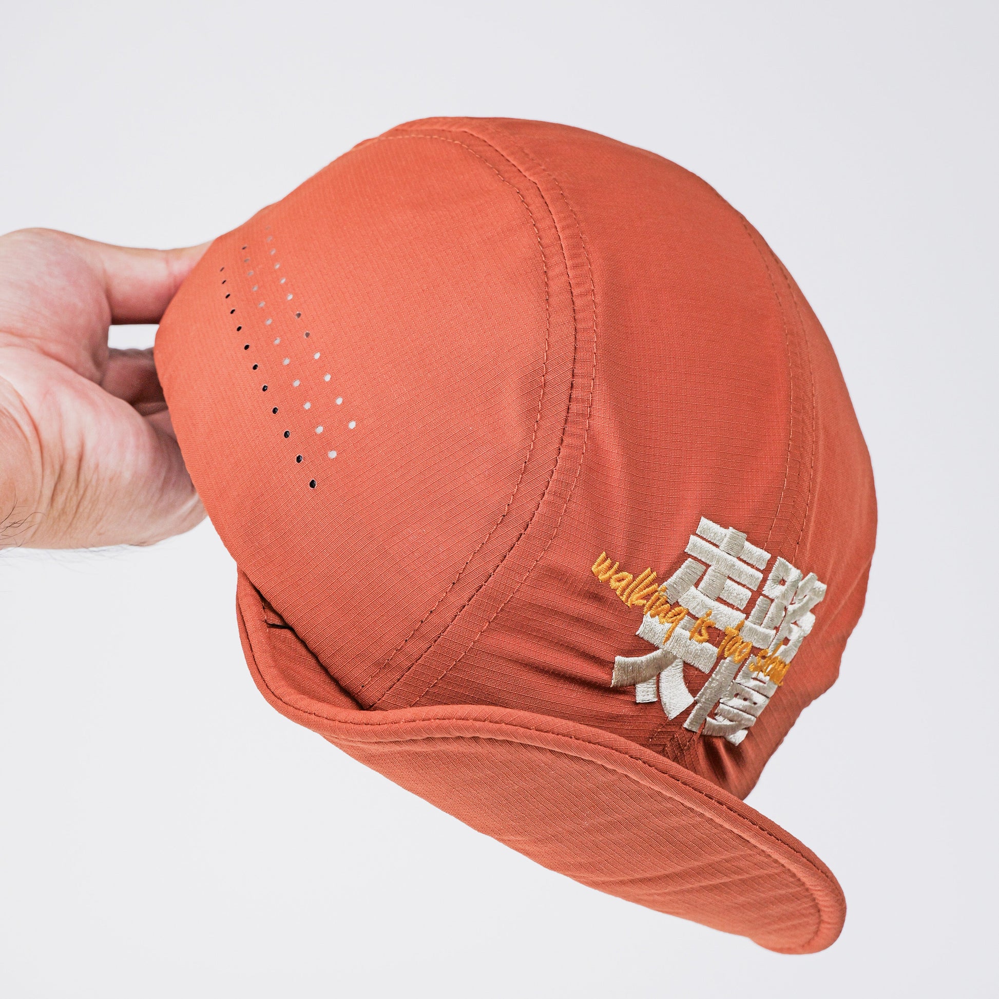 A hand holding a terracotta orange baseball cap with a white and gold embroidered "走路太慢 walking is too slow"logo on the side, set against a plain light gray background.