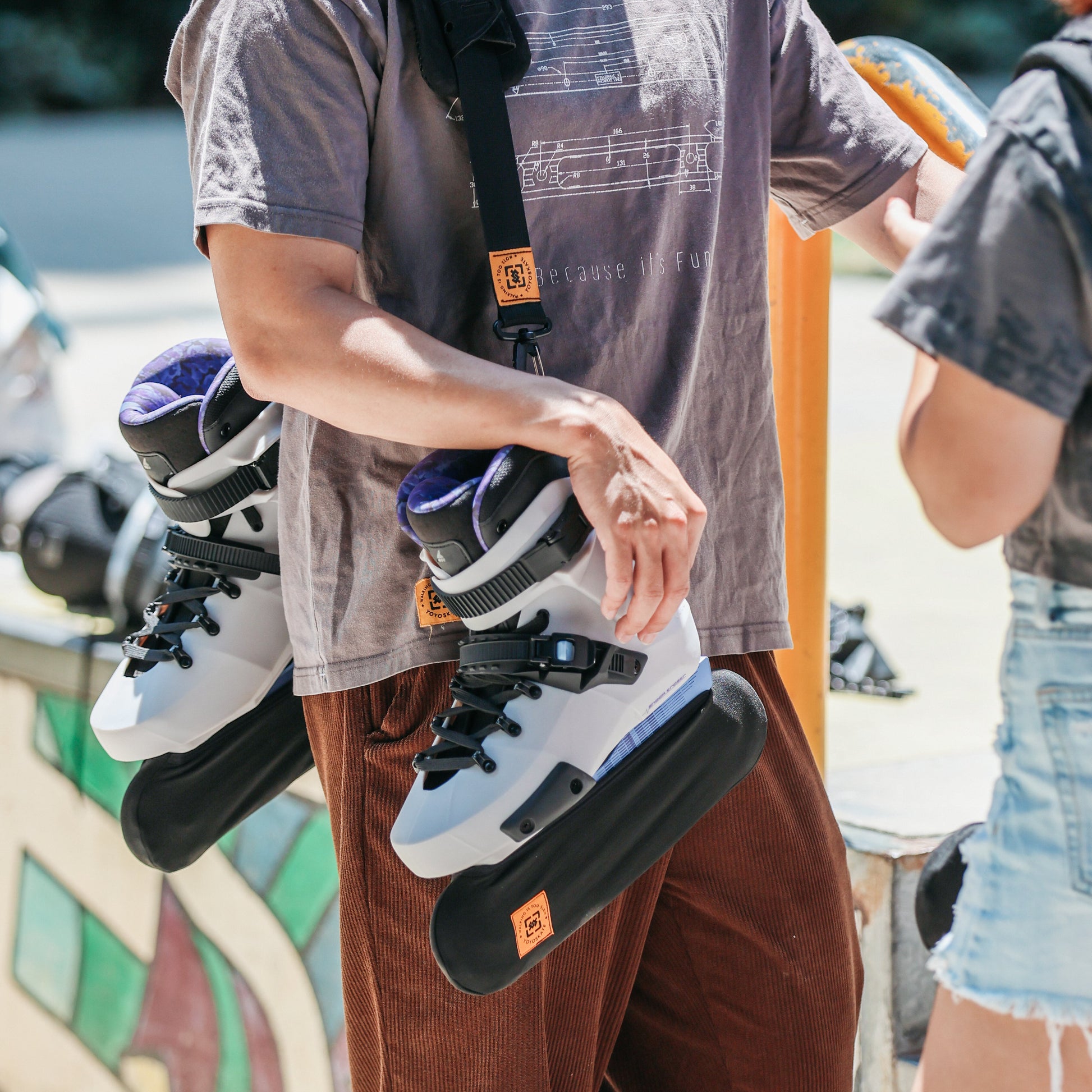 A person outdoors wearing a gray graphic T-shirt and brown pants, carrying a black strap-mounted inline skate across their shoulder and holding a second skate in one hand. Another person is partially visible at the right edge.
